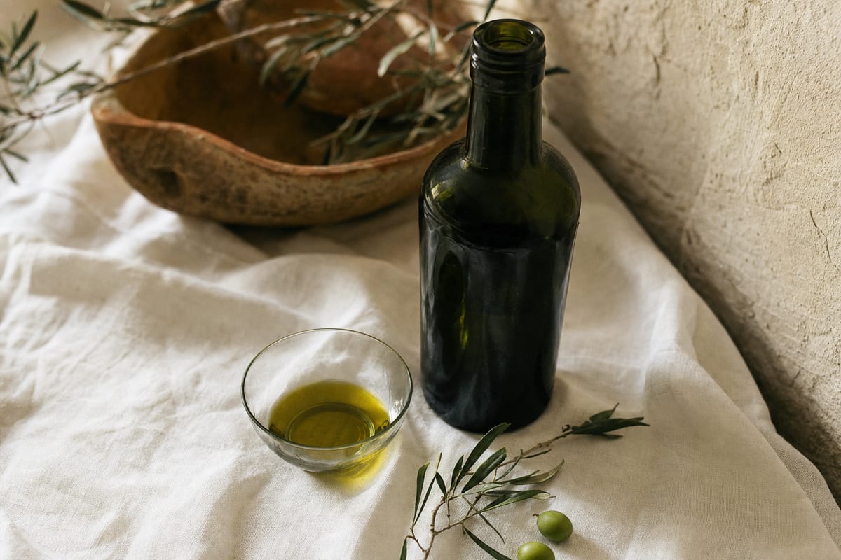 Botella de aceite de oliva virgen extra en vidrio verde oscuro junto a un vaso de cata con aceite y hojas de olivo, sobre lino claro